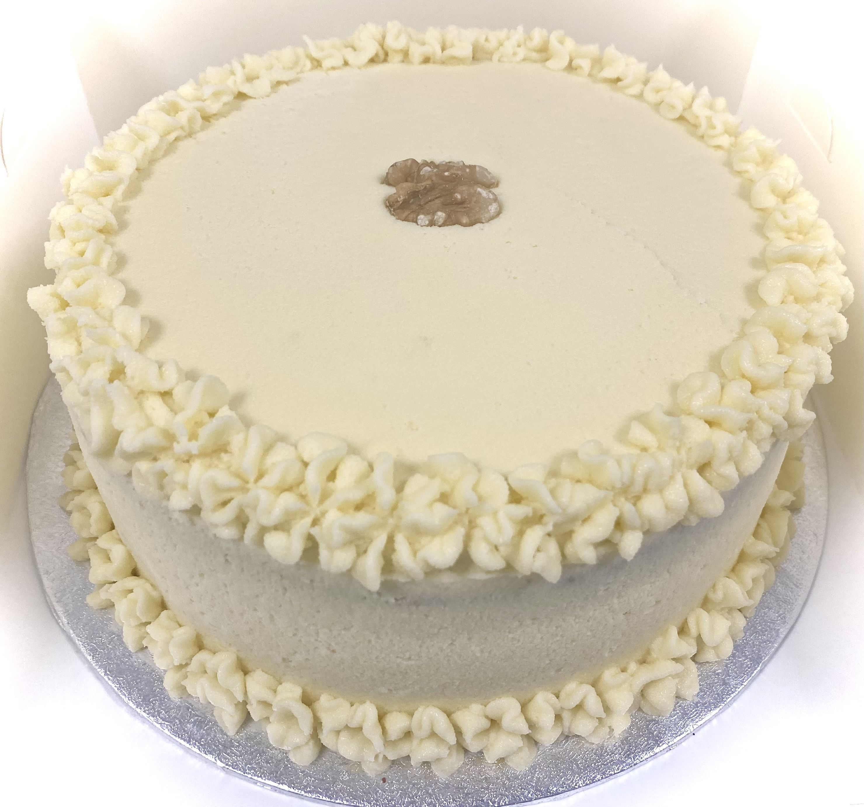 A round, award-winning carrot cake with Philadelphia cream cheese frosting, featuring decorative piped borders and a walnut piece placed at the center on top. The cake is set on a silver board.